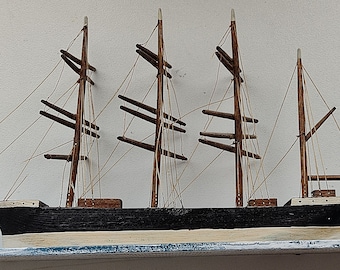 May include: A wooden model ship with three masts and a black hull. The ship has a white base and is on a white shelf.