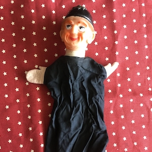 Puede incluir: Un títere de mano vintage con una túnica de fieltro negro y una cara de fieltro blanco. El títere lleva un sombrero negro y tiene una expresión sonriente.