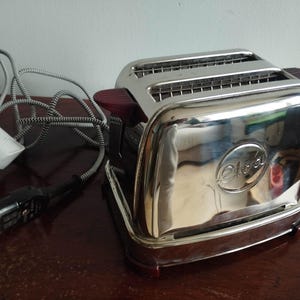 Könnte beinhalten: Ein verchromter Vintage-Toaster mit einem dunkelholzfarbenen Griff und Sockel. Der Toaster hat zwei Schlitze und ein Netzkabel mit weißem Stecker. Der Markenname "Oloa" ist auf der Vorderseite eingeprägt. Der Toaster steht auf einer dunklen Holzoberfläche.