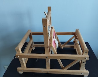May include: A wooden loom with two spools and a red and white woven piece in the center. The loom is made of natural wood and has a simple design.