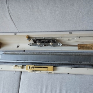 Può includere: Una macchina da maglia bianca e grigia con un telaio in metallo e una custodia in plastica bianca. La macchina è aperta e gli aghi sono visibili. La macchina è in posizione chiusa e la custodia è chiusa.