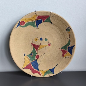 May include: A round, beige ceramic plate with a textured surface featuring hand-painted abstract shapes in red, yellow, green, and blue. The design includes thin black lines connecting the geometric forms, with the signature "Bellen" in black ink.