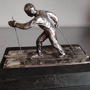 May include: A silver-colored sculpture of a skier in motion, poised with ski poles, atop a rectangular base. The skier is depicted in a dynamic pose, suggesting speed and action. The base is a dark, rectangular block.