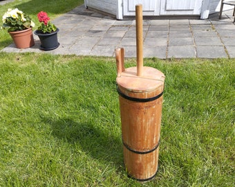 May include: A wooden butter churn with a wooden handle and a black metal band around the top. The churn is sitting on a green grassy surface.
