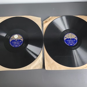 Könnte beinhalten: Zwei schwarze Schallplatten mit blauen und goldenen Decca-Etiketten, jeweils in einer hellbraunen Papierhülle. Die Etiketten tragen den Text Decca The Supreme Record, mit spezifischen Titelinformationen in weißer Schrift auf blauem Grund.