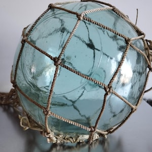 Puede incluir: Un flotador de vidrio transparente, grande y redondo, con un tinte azul claro, envuelto en una red de cuerda marrón. El flotador tiene una superficie texturizada y está diseñado para ser flotante. La red de cuerda está anudada y tiene una borla en el extremo.