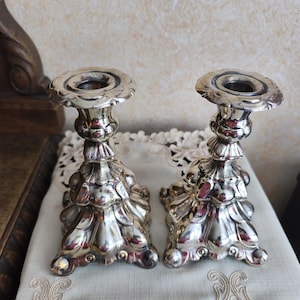 Può includere: Due candelabri argentati ornati con un motivo floreale. I candelabri hanno una base ampia e una superficie strutturata dettagliata. I portacandele sono posizionati su un tessuto bianco.