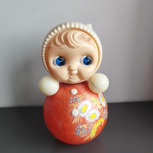 May include: A vintage roly-poly toy with a doll face, blue eyes, and a bonnet. The red body is decorated with a floral design. It has two white spheres on the sides and is designed to rock. A classic children's toy.