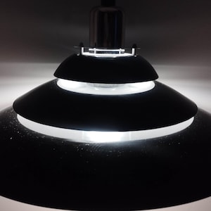 May include: A black, tiered pendant light fixture with a bright white light emanating from the center. The fixture has a metallic silver top and a series of black, circular shades. The light is suspended from the ceiling.