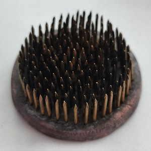 Op de afbeelding: Een ronde, donkerbruine bloemenspin met talrijke messing pinnen. De pinnen zijn dicht op elkaar geplaatst en vormen een textuurrijk oppervlak. De basis heeft een verweerde uitstraling, met een diameter van ongeveer 7 cm.