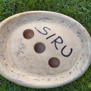May include: A wooden, oval-shaped object with three holes and the word "SIRU" written on it. The object has a light brown color and a rough texture. The number "27156" is written on the object.