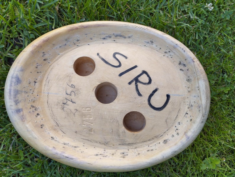 May include: A wooden, oval-shaped object with three holes and the word "SIRU" written on it. The object has a light brown color and a rough texture. The number "27156" is written on the object.