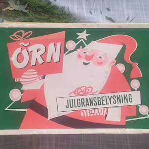 May include: A vintage Christmas light box with a green background and a red and white illustration of Santa Claus holding a sign that says "ÖRN JULGRANSBELYSNING".