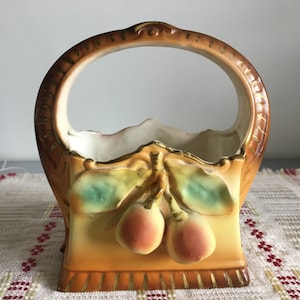Könnte beinhalten: Ein Keramik-Obstkorb mit einem braunen und goldenen Griff und einem weißen Innenraum. Der Korb ist mit bemalten grünen Blättern und zwei roten Kirschen verziert.