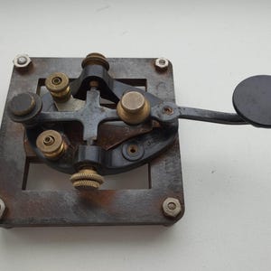 May include: An antique Morse code telegraph key. The device features a square, rusty metal base with a complex arrangement of levers, screws, and a black circular key. The key is a vintage communication tool.