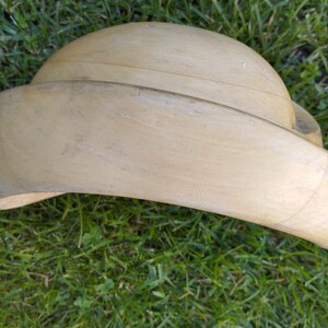 May include: A light brown wooden hat form, used for shaping hats. The form is curved and has a wide brim.