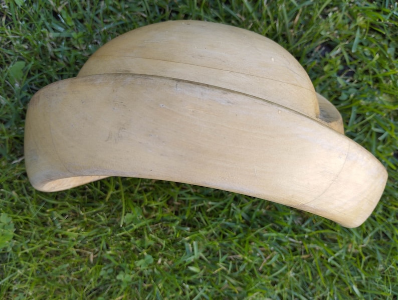 May include: A light brown wooden hat form, used for shaping hats. The form is curved and has a wide brim.