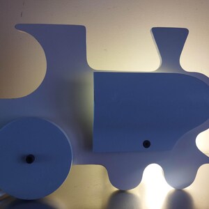 May include: A white train-shaped wall lamp with a smooth silhouette, featuring a large circular wheel detail on the side, a central rectangular section, and a chimney-like top. The lamp emits a soft, warm glow from behind its structure.