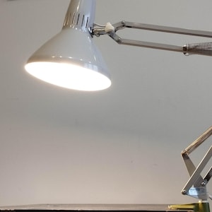 Puede incluir: Una lámpara de escritorio ajustable de plata con una pantalla blanca. La lámpara está encendida y la luz brilla.