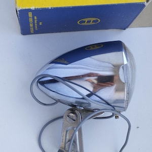May include: A vintage chrome Bosch bicycle headlight with a blue top section featuring the Bosch logo. The headlight includes a metal mounting bracket and grey electrical wiring. It is positioned in front of its original yellow and blue cardboard box.