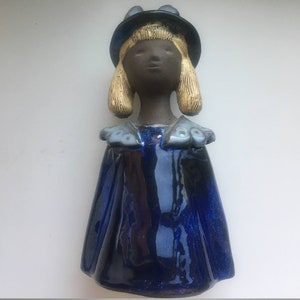 以下が含まれることがあります： 暗い肌、金髪、青い帽子とケープを着た女の子の陶器の置物。 置物は光沢のある仕上がりで、白い背景に配置されています。 ケープにはフリルの襟が付いています。