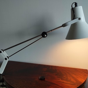 Puede incluir: Una lámpara de escritorio blanca con una pantalla cónica y un brazo ajustable. La lámpara está montada sobre una superficie de madera oscura. El brazo de la lámpara es negro y blanco, y la pantalla está inclinada hacia abajo, proyectando una luz cálida.