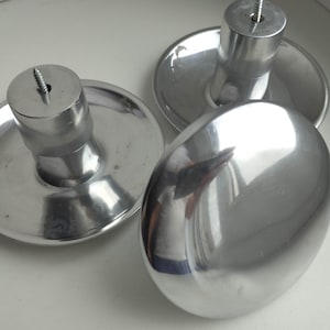 Puede incluir: Tres pomos de metal plateado pulido con una superficie superior lisa y redondeada y una base cilíndrica. Cada pomo presenta un tornillo de metal roscado que sobresale del centro de la parte superior, diseñado para fijarse a muebles.