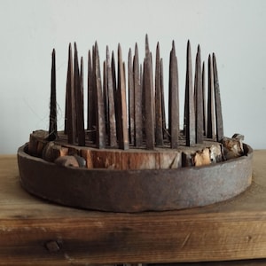 May include: A vintage, circular flower frog with numerous metal spikes. The spikes are arranged in a dense pattern, rising from a wooden base encased in a rusty metal ring. The piece sits on a wooden surface, showcasing its aged appearance.