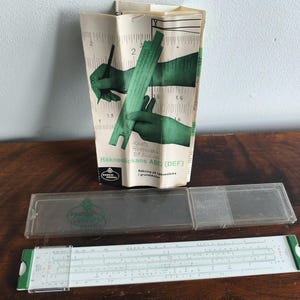 Pode incluir: Uma régua de cálculo branca e verde com um estojo de plástico transparente e um manual de instruções em papel. O manual apresenta ilustrações verdes de mãos usando a ferramenta e inclui o texto "Räknestickans ABC (DEF)" e "Faber-Castell".