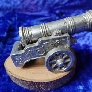 Puede incluir: Un cañón plateado en miniatura detallado, con detalles intrincados, que incluyen un cañón texturizado y tallas decorativas. El cañón descansa sobre una pequeña base de madera redonda, sobre un fondo azul oscuro.