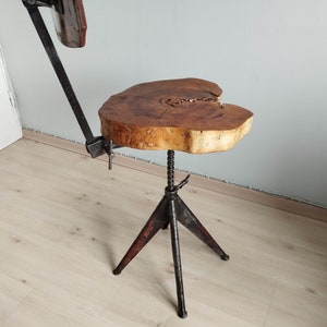 May include: A rustic wooden table top with a natural edge sits on a black metal adjustable tripod base. The table top is made of a light brown wood with a natural grain pattern. The base is made of black metal with a distressed finish.