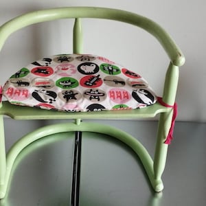 May include: A light green wooden chair with a curved back and a seat cushion featuring a colorful pattern of black cats, red and white polka dots, and the word "Aha!" repeated. The chair has red ribbon ties on the back.