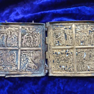 Puede incluir: Una cubierta de libro antigua y abierta con detallados grabados en relieve. La cubierta metálica está dividida en cuatro secciones a cada lado, que representan escenas religiosas. El metal tiene un aspecto deslustrado y envejecido, con un fondo de terciopelo azul.