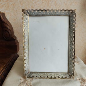 Könnte beinhalten: Ein rechteckiger, verzierter Bilderrahmen mit gold- und silberfarbenem Finish. Der Rahmen hat ein filigranes, spitzenartiges Design an den Rändern und einen weißen Innenraum. Er steht auf einer cremefarbenen Oberfläche.