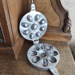 May include: An open, antique silver-colored cookie iron with a hinged design. The top half has oval molds, while the bottom half features a star-shaped center and striped molds. The handle is attached to the top half.