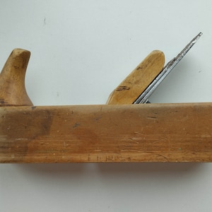 Puede incluir: Un cepillo de mano de madera vintage con una cuchilla de metal. El cepillo es de madera de color marrón claro y tiene un mango curvo. La cuchilla de metal es visible en el centro del cepillo. La herramienta parece estar bien usada y muestra signos de desgaste.