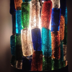 Puede incluir: Una lámpara decorativa con forma cilíndrica, compuesta por piezas de vidrio coloridas y texturizadas. El vidrio está dispuesto en un patrón de mosaico, con tonos de azul, verde, naranja y transparente. La lámpara emite un brillo cálido, resaltando los variados colores y texturas del vidrio.