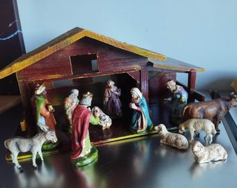 May include: A wooden nativity set featuring a dark red stable structure with a peaked roof. The set includes hand-painted figurines of figures in robes, a baby in a manger, a donkey, a brown cow, and three sheep arranged in front of the stable.