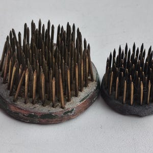 Op de afbeelding: Twee vintage bloemenkikkers, ook wel kenzans genoemd, voor bloemschikken. Eén is groter, met een ronde basis en een mix van gouden en donkere metalen pinnen. De kleinere heeft een donkere basis en donkere metalen pinnen.