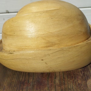 May include: A wooden hat form, also known as a millinery block, is made of light brown wood and has a rounded, bowl-like shape.