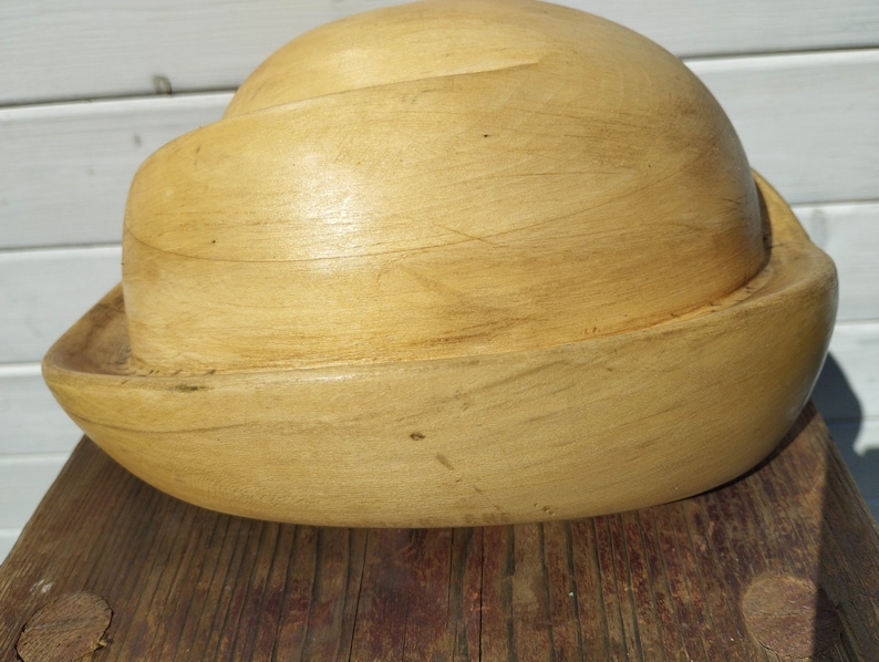 May include: A wooden hat form, also known as a millinery block, is made of light brown wood and has a rounded, bowl-like shape.