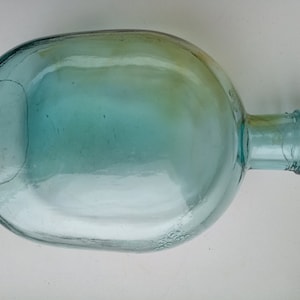 May include: A clear glass bottle with a rounded body and a long neck. The bottle is light blue in color and has a slight green tint.