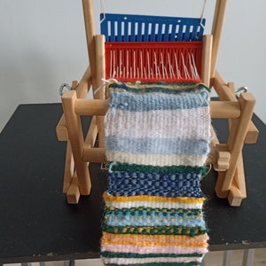 Könnte beinhalten: Ein Holzwebstuhl mit einem bunten gewebten Wandteppich, der daran hängt. Der Wandteppich ist aus verschiedenen farbigen Garnen gefertigt und hat ein gestreiftes Muster.