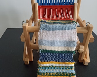 Op de afbeelding: Een houten weefgetouw met een kleurrijk geweven wandkleed dat er vanaf hangt. Het wandkleed is gemaakt van verschillende gekleurde garens en heeft een gestreept patroon.