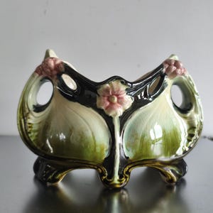 Può includere: Un vaso in ceramica con finitura lucida nei toni del verde bosco profondo, crema e rosa tenue. Il contenitore presenta una forma organica curva con due aperture laterali circolari, un rilievo floreale rosa centrale e quattro piedini.