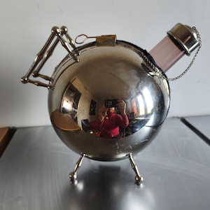 Puede incluir: Una cubitera esférica plateada con tapa abisagrada y tres patas pequeñas. Se ve un cilindro de vidrio rosa, sujeto por una tapa y cadena plateadas. La superficie refleja el entorno.