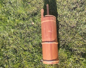 May include: A tall, cylindrical wooden butter churn with a handle and a lid. The churn is made of light brown wood with darker brown bands and accents. The churn is sitting on a bed of green grass.