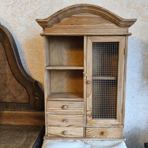 Może przedstawiać: Jasna drewniana szafka z zakrzywionym blatem, półkami, szufladami i drzwiami z siatki drucianej. Szafka posiada trzy małe szuflady z okrągłymi drewnianymi gałkami oraz pojedyncze drzwi z wkładką z siatki drucianej. Szafka wykonana jest z jasnego drewna.