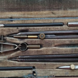May include: A vintage compass set in a wooden case. The set includes three compasses, a ruler, and a pencil. The compasses are made of metal and have a brass finish. The ruler is made of wood and has a brass edge. The pencil is made of wood and has a brass tip.