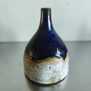 May include: A handcrafted ceramic vase with a dark blue glazed top and a textured, off-white base. The vase has a unique shape, with a narrow neck and a rounded body. The base has a gold band separating it from the blue top.